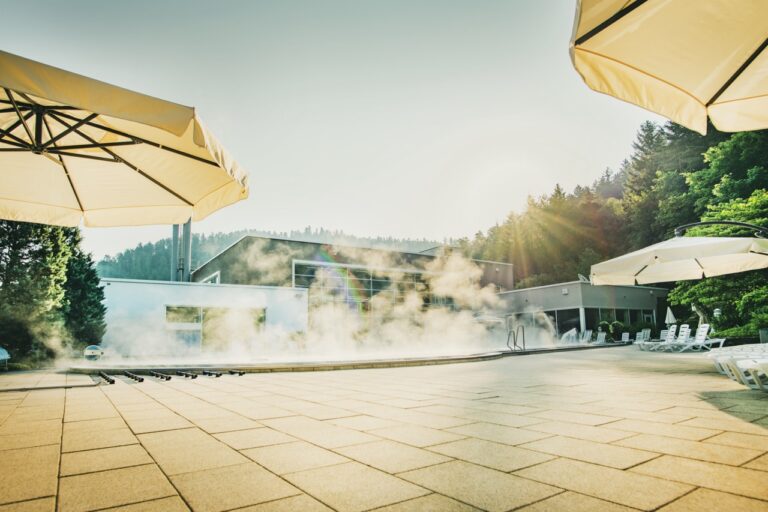 Sonnenterrasse eines Thermalbads mit Dampfschwaden, Sonnenschirme, Wald und moderne Gebäude im Hintergrund.