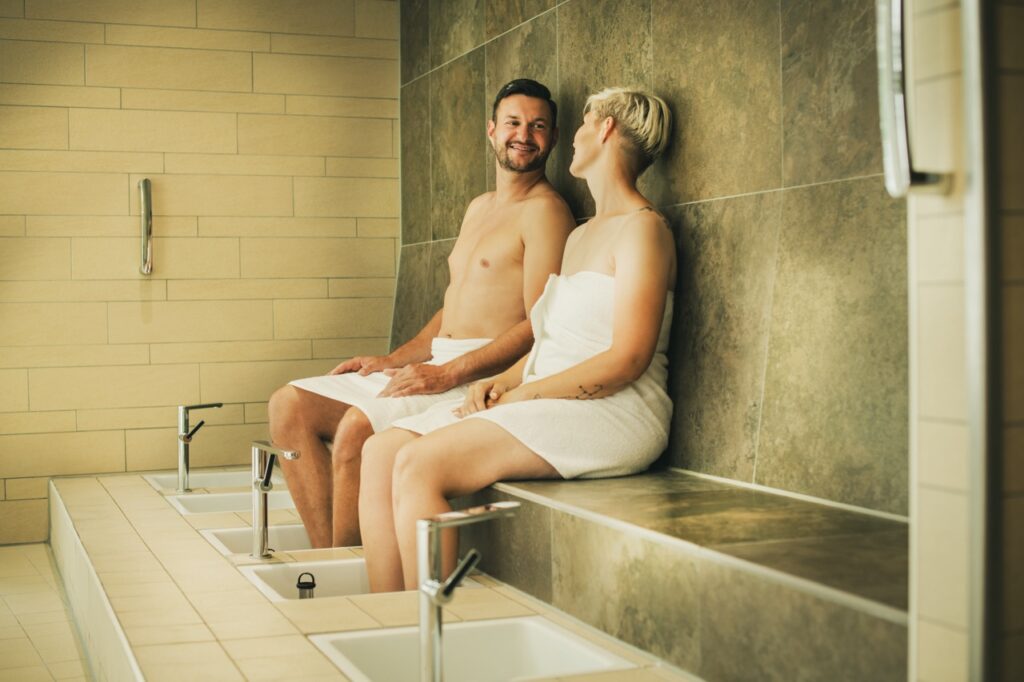 Deux personnes avec des serviettes dans un espace spa, assises sur un banc en pierre, avec des bains de pieds.