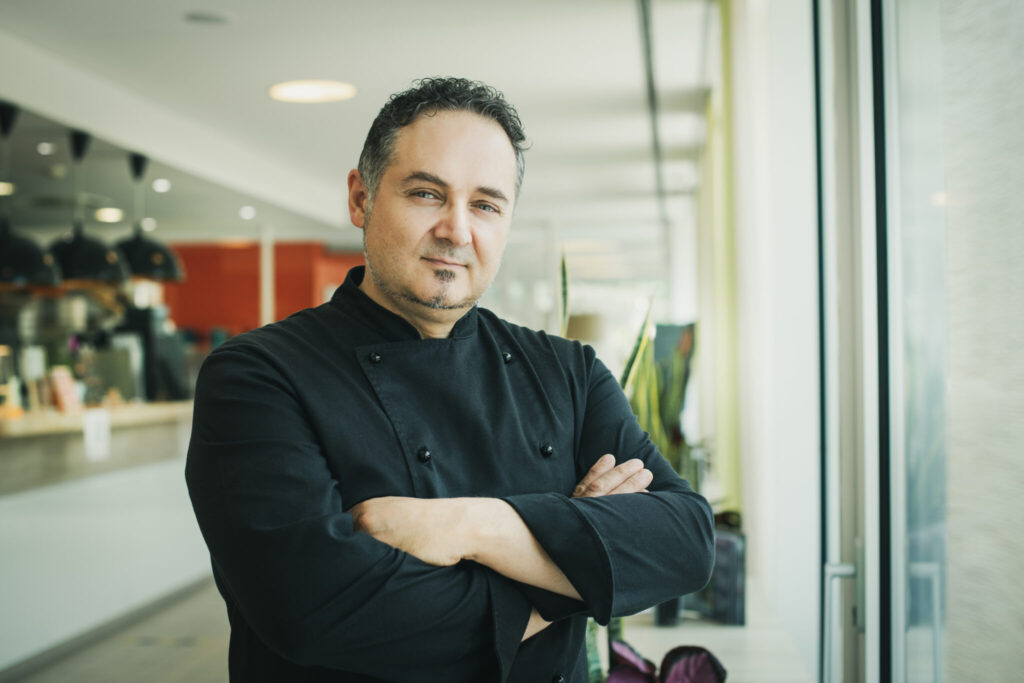 Un homme d'âge moyen se tient debout, les bras croisés, dans un restaurant ou un café moderne et lumineux. Il porte une veste de cuisine noire et regarde la caméra avec un léger sourire et une expression confiante. En arrière-plan, on distingue vaguement des tables, des plantes et des éléments de cuisine, ce qui indique un environnement gastronomique professionnel.